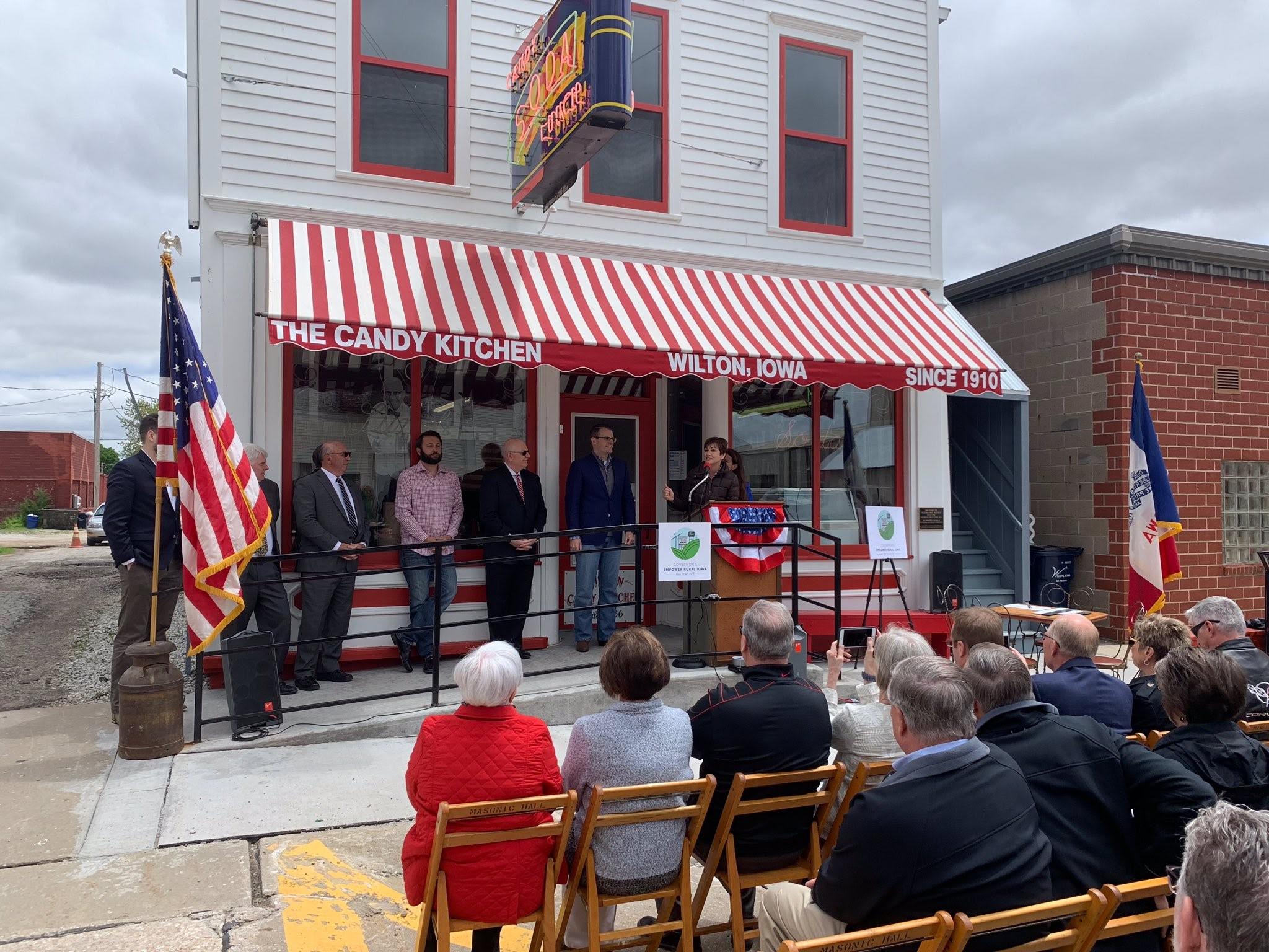 A small crowd watches a press conference featuring Governor Kim Reynolds on the storefront of The Candy Kitchen in Wilton, Iowa.